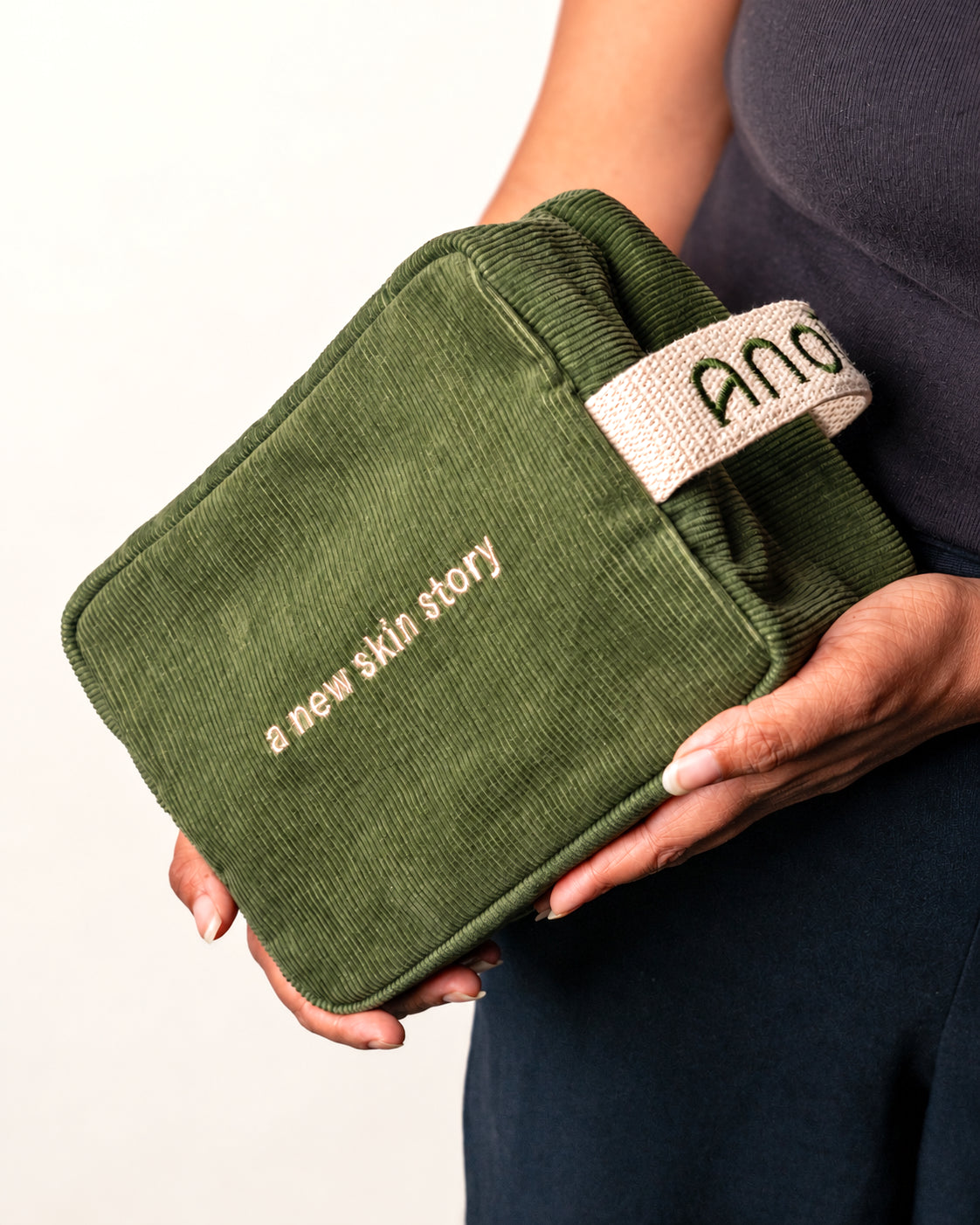 Green Anoū Carryall corduroy travel pouch held in two hands, with embroidered “a new skin story” wording and an Anoū side tab.